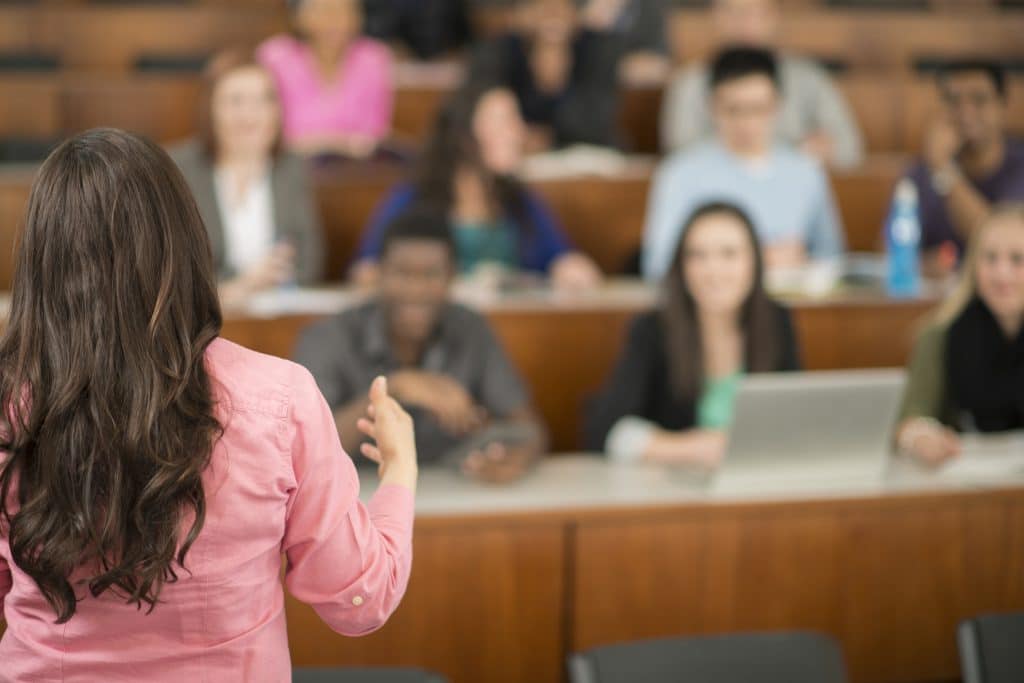 A multi-ethnic group of college age students are sitting in a row in a lecture hall and are listening to their professor.
