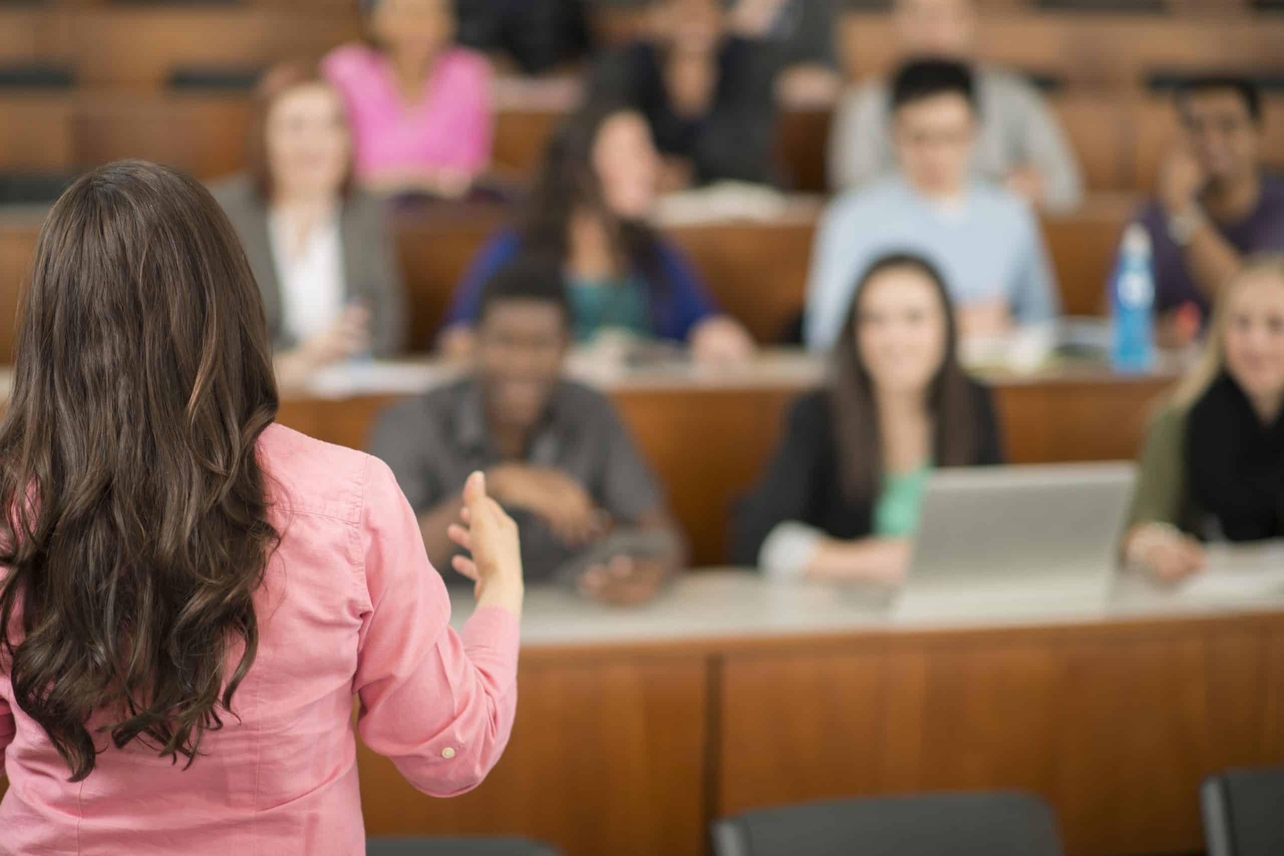 A multi-ethnic group of college age students are sitting in a row in a lecture hall and are listening to their professor.